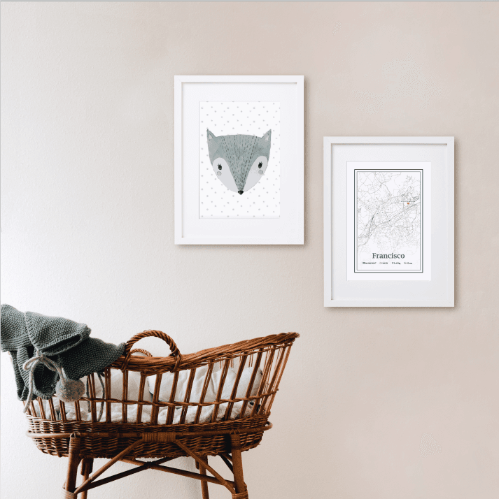 Dois quadros brancos com ilustrações pendurados numa parede bege acima de um berço de verga castanho com manta cinzenta.